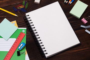 Empty spiral notebook with white page and multicolored stationery on brown wooden table. Workplace...