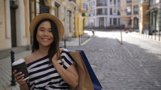 Rear view of asian elegant hipster girl in striped shirt and hat carrying shopping bags and coffee cup in hands walking down the street after shopping. Beautiful joyful female looking back and winking