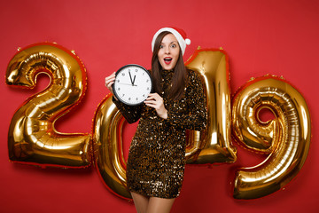 Merry Santa girl in shiny glitter dress, Christmas hat hold round clock isolated on bright red wall background, golden numbers air balloons studio portrait. Happy New Year 2019 holiday party concept.