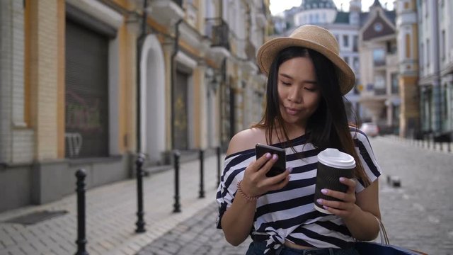 Carefree asian hipster girl in trendy clothes taking photos on smart phone while walking along city street. Positive traveller young woman with shopping bags and coffee to go enjoying summer day.