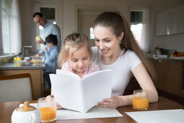 Happy family with little kids spend free time together at new home. Smart preschool adorable daughter sits on attractive young mother lap looking reading interesting book, father and son on background