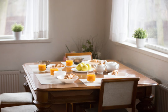 Fresh Healthy Breakfast Served At The Dinner Table In Modern Cozy Kitchen Daylight Through The Windows, Empty Chairs No People. Orange Juice And Pastries Croissants Prepared For Family In The Morning