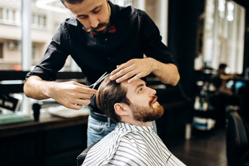 Barber with mustache dressed in a black shirt with a red bow tie makes a stylish hairstyle to young...