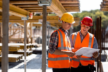 Two civil engineers dressed in orange work vests and helmets explore construction documentation on...