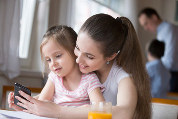 Woman have fun together with little adorable girl sitting indoors. Mother and daughter looking at mobile phone screen watching cartoons making video call or playing games father with son on background