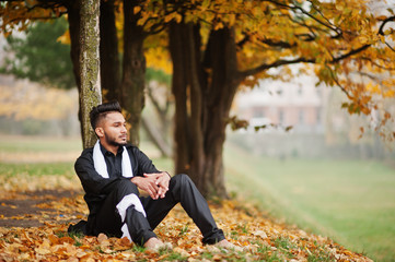 Indian stylish man in black traditional clothes with white scarf posed outdoor against yellow autumn leaves tree.