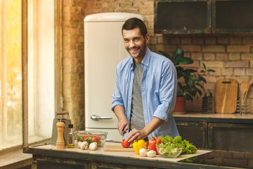 It's so delicious! Casual happy young man preparing salad at home in loft kitchen and smiling,...