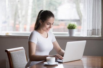 Attractive millennial young woman sitting in kitchen at table looking at screen typing chatting using laptop alone at home. Smiling female communicate online with friend feels good satisfied and glad