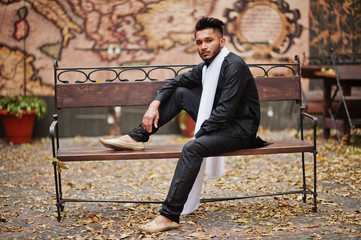 Indian stylish man in black traditional clothes with white scarf posed outdoor sitting on bench.