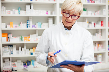 Serious mature female pharmacist filling documents on a clipboard. Shelves with medications in the background. Medicine and healthcare concept
