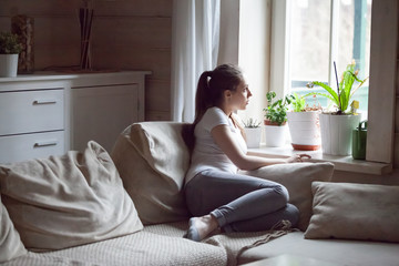 Pensive attractive millennial woman sitting alone on sofa in living room at home thinking...