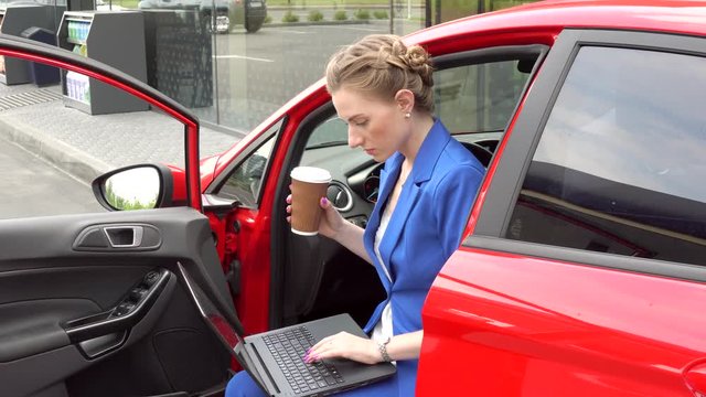 Busy woman sits in car and works. She holds laptop on nap. Also girl has cup of coffee in hands. She is calm and concentrated.
