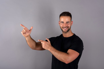 A handsome young man in a black tshirt pointing with his fingers in front of a grey background in the studio.