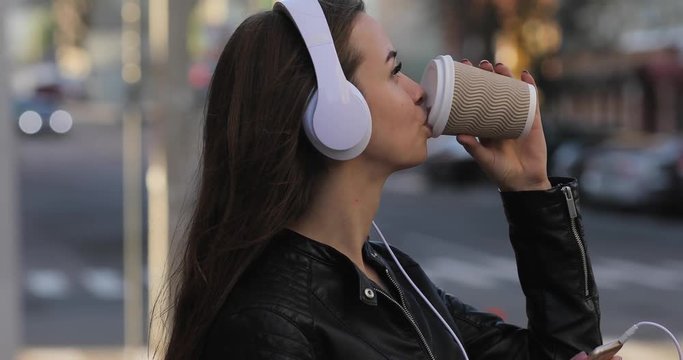 Modern woman with coffee cup in city listening to music in headphones, lifestyle