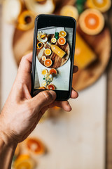 male hands are taking pictures of orange juice still life in a glass bottle lying on a dark wooden plate on a wooden table with orange slices flatlay