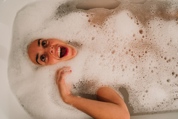 .Young and funny woman taking a bath of foam making bubbles in the white bath of her house. A fun time with joyful gestures. Lifestyle.