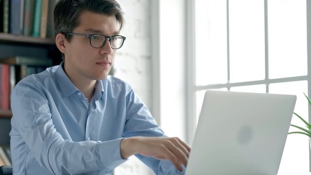 Young beautiful smiling student man is sitting at desk in big spacious library with pile of books. She is writing something in copybook, laptop is in front of her