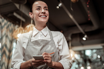 Funny joke. Happy cafe worker wearing apron and making notes in her copybook