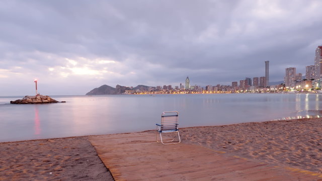 Playa de poniente desierta al atardecer en Benidorm, con el fato y las luces de los edificios en un d&iacute;a nublado con una silla abandonada en el camino de madera en la orilla