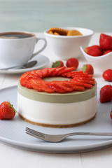 Cooking demo. No-bake two-layered mini strawberry matcha cheesecake. Strawberry slices decorate the top of the cake. 3 cups with coffee, cookies and strawberry in background.