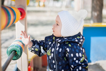 Side portrait of a little toddler boy in woolen hat and jumpsuit playing with a toy abacus at playground in a sunny cold day. Sustainable family finance concept