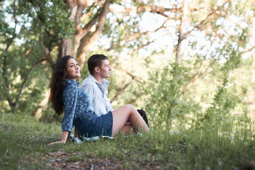 Naklejka na ściany i meble young couple sitting on the grass in the forest and looking on sunset, summer nature, bright sunlight, shadows and green leaves, romantic feelings