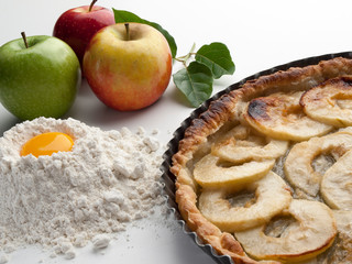 Pastel de manzana en su molde horneado junto a manzanas crudas, harina y huevo, vistos frontalmente