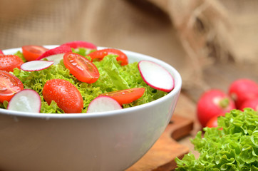 Close-up of green salad Lollo Biondo with tomatoes and radishes in a white bowl on wooden table, a pitcher of oil in the background - vertical photo