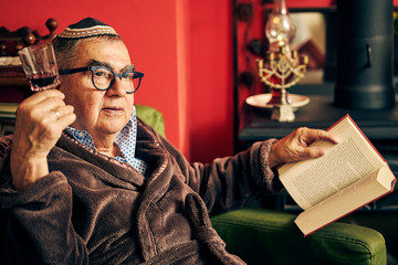 Jewish senior with glasses sitting in the armchair reading a torah book and drinking kosher wine