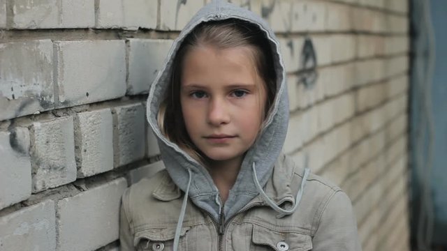 Sad young girl looking at camera with pain and reproach against grey brick wall. Difficult child wearing hooded jacket close up portrait outdoors.