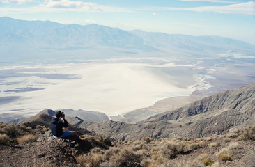 Fototapeta premium A photographer / videographer is sitting on the edge of the high rock overseeing and taking pictures of the fantastic landscape of Death Valley / USA