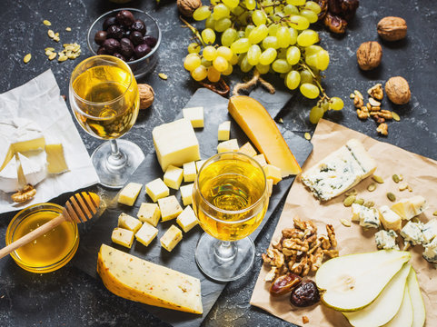 Different types of cheese on board, olive, fruits, almond and wine glasses on stone table
