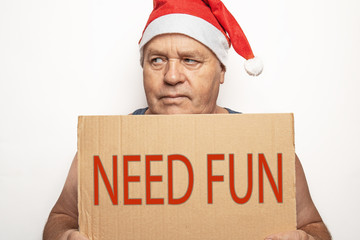 Funny upset and angry mature man in red Christmas Santa hat holds cardboard sign with inscription - need fun on white background