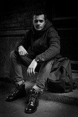 A young man with a hipster hanging on an outer garment on the stairs. Hoodshavy man in a wool jacket and high quality slippers with a backpack. Conceptual black and white photo.