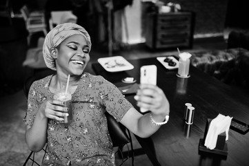 Stylish african woman in red shirt and hat posed indoor cafe, drinking strawberry lemonade and making selfie on phone.