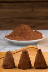 Three chocolate truffles on a wooden background, cocoa powder for sprinkling in a round plate.
