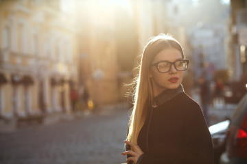 Outdoor portrait of gorgeous blonde woman with red lips wearing glasses and coat, posing at the old sunny street.  Empty space