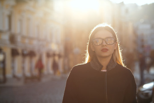 Outdoor portrait of stylish blonde girl with red lips wearing glasses coat, posing at the old sunny street. Empty space