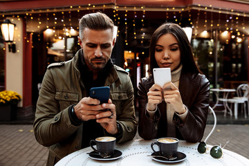 Cafe. Couple. Technology. Man and woman in warm casual clothes are using their smartphones while sitting in the cafe outdoors