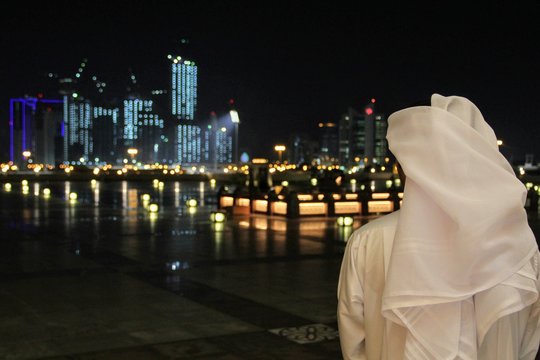 An Arab Qatari man wearing a white thobe or thwab (Arab garment) and white ghutra (or keffiyeh or headress) looking out to the evening or night skyline of Doha, Qatar
