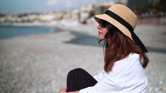 Side view of tourist woman with long brown hair wearing hat and sunglasses sitting on pebble beach and looking at sea. Locked down real time medium shot