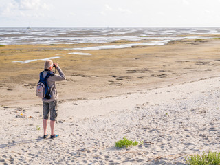 Senior man with binoculars looking at tidal flats at low tide of Waddensea from beach of Boschplaat...
