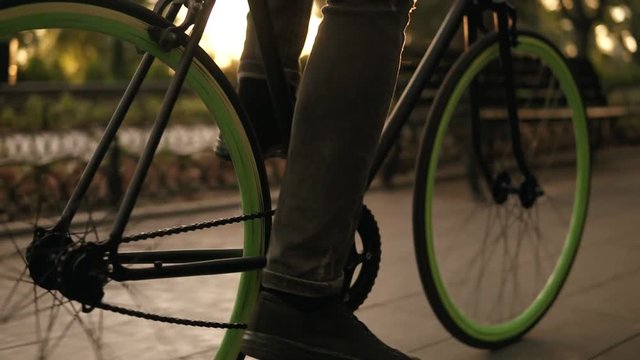 Close up footage of male feet cycling a bicycle in the morning park by paved road. Side view of a young man riding a trekking bike with green wheels, wearing black sneakers. Lens flares on the