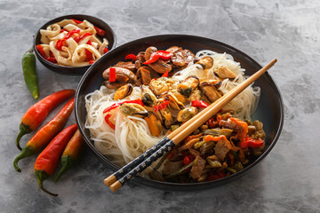 Rice noodles with seafood, salad, fried mushrooms and red pepper stand on a gray table.