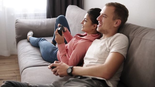 Relaxed young couple, playing video games at home while lying on sofa and enjoying themselves. They have great weekend indoors playing tv game with joysticks. Side view