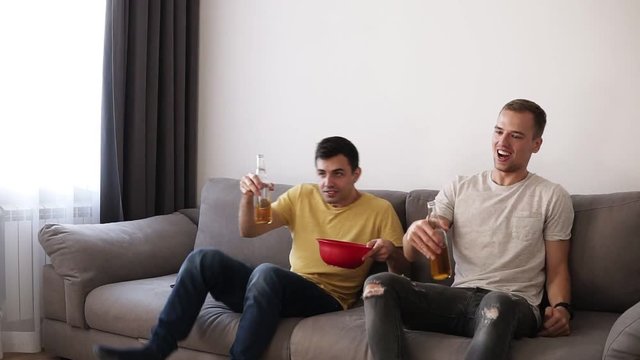 Picture of two friends come to the living room watching tv sitting in a comfortable sofa at home, eating chips from the bowl and drinking beer from the bottles, enjoying their leisure time
