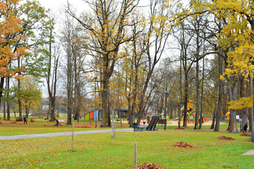 Autumn park landscape with old trees, playground for children and workers raking fallen autumn leaves and cleaning public city recreation park in early autumn morning.