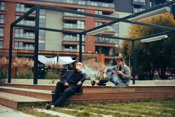 Two friends sit in the park on a bench. Friendship. Two fashion men in coats  . Man vape, his friend uses the phone