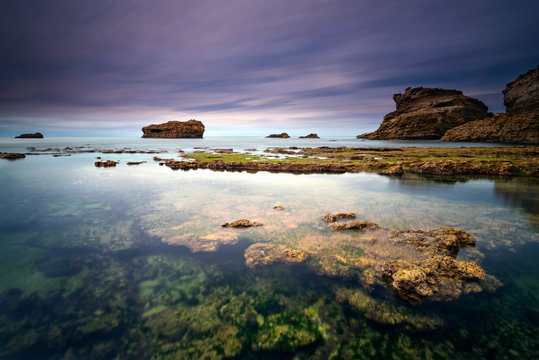 Vue sur un ensemble de rochers en pose longue depuis la plage avec en premier plan un bassin transparent
