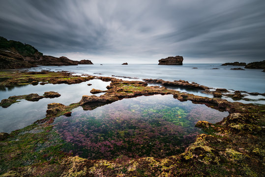 Vue sur un rocher en pose longue depuis la plage avec en premier plan un bassin translucide